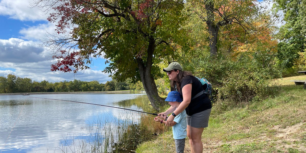Mother leaning over her son helping him hold a fishing rod on a lake shore